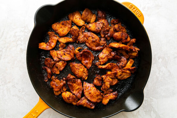 How to make chicken fajita burritos, step 3: Cook the chicken fajitas. Sliced chicken marinated in spices fills a Yellow Staub cast iron skillet. The chicken has been cooked and browned. The skillet sits atop a creamy white textured surface.