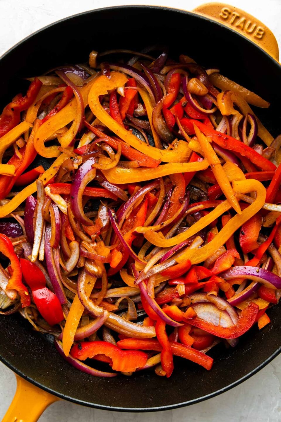 How to make chicken fajita burritos, step 4: Cook the fajita veggies. Sliced bell Peppers. & onions fill a Yellow Staub cast iron skillet. The fajita veggies have been cooked in oil & seasoned with kosher salt and ground black pepper. The skillet sits atop a creamy white textured surface.