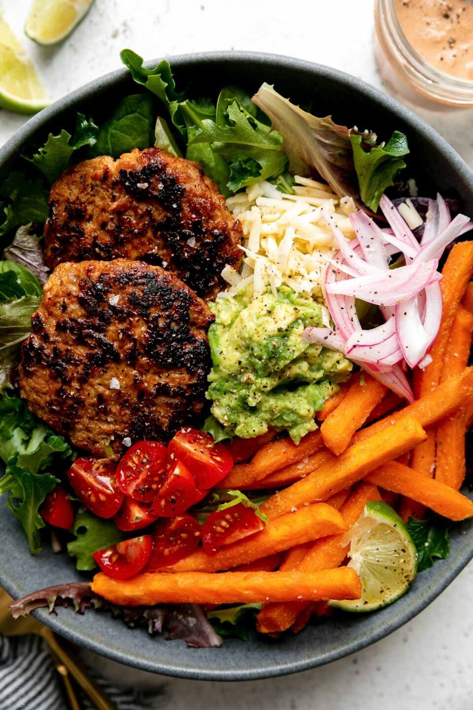 An assembled serving of Chipotle Turkey Burger Bowls with Sweet Potato Fries fills a blue ceramic bowl. The turkey burgers are nestled atop a bed of greens with baked Alexia Sweet Potato Fries, smashed avocado, pickled red onions, quartered cherry tomatoes, shredded cheese and a lime wedge. The bowl sits atop a creamy white textured surface with a small jar of chipotle lime crema, two lime wedges, a blue and white striped linen napkin and a set of gold silverware surrounding the bowl.