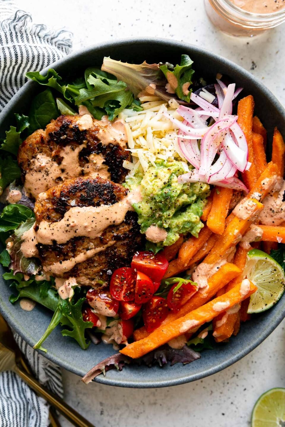 An assembled serving of Chipotle Turkey Burger Bowls with Sweet Potato Fries fills a blue ceramic bowl. The turkey burgers are nestled atop a bed of greens with baked Alexia Sweet Potato Fries, smashed avocado, pickled red onions, quartered cherry tomatoes, shredded cheese, a lime wedge and chipotle lime crema drizzled over top. The bowl sits atop a creamy white textured surface with a small jar of chipotle lime crema, two lime wedges, a blue and white striped linen napkin and a set of gold silverware surrounding the bowl.