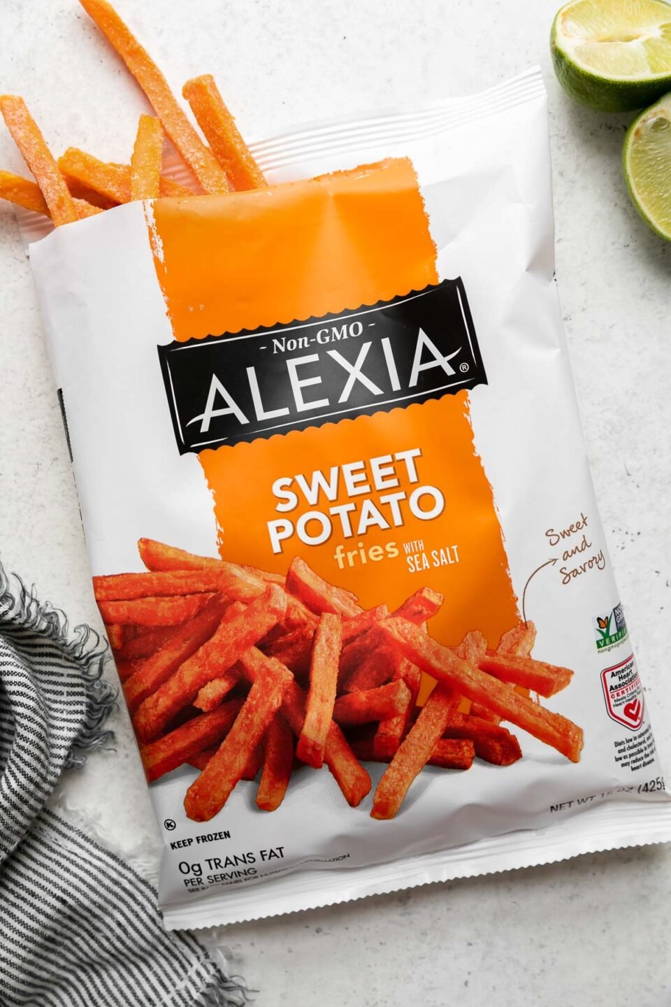 A single bag of Alexia Sweet Potato Fries with Sea Salt rest atop a creamy white textured surface. The bag has been cut open and a handful of frozen sweet potato fries peek up out of the bag. Two lime halves and a blue and white striped linen napkin surround the bag of frozen sweet potato fries.
