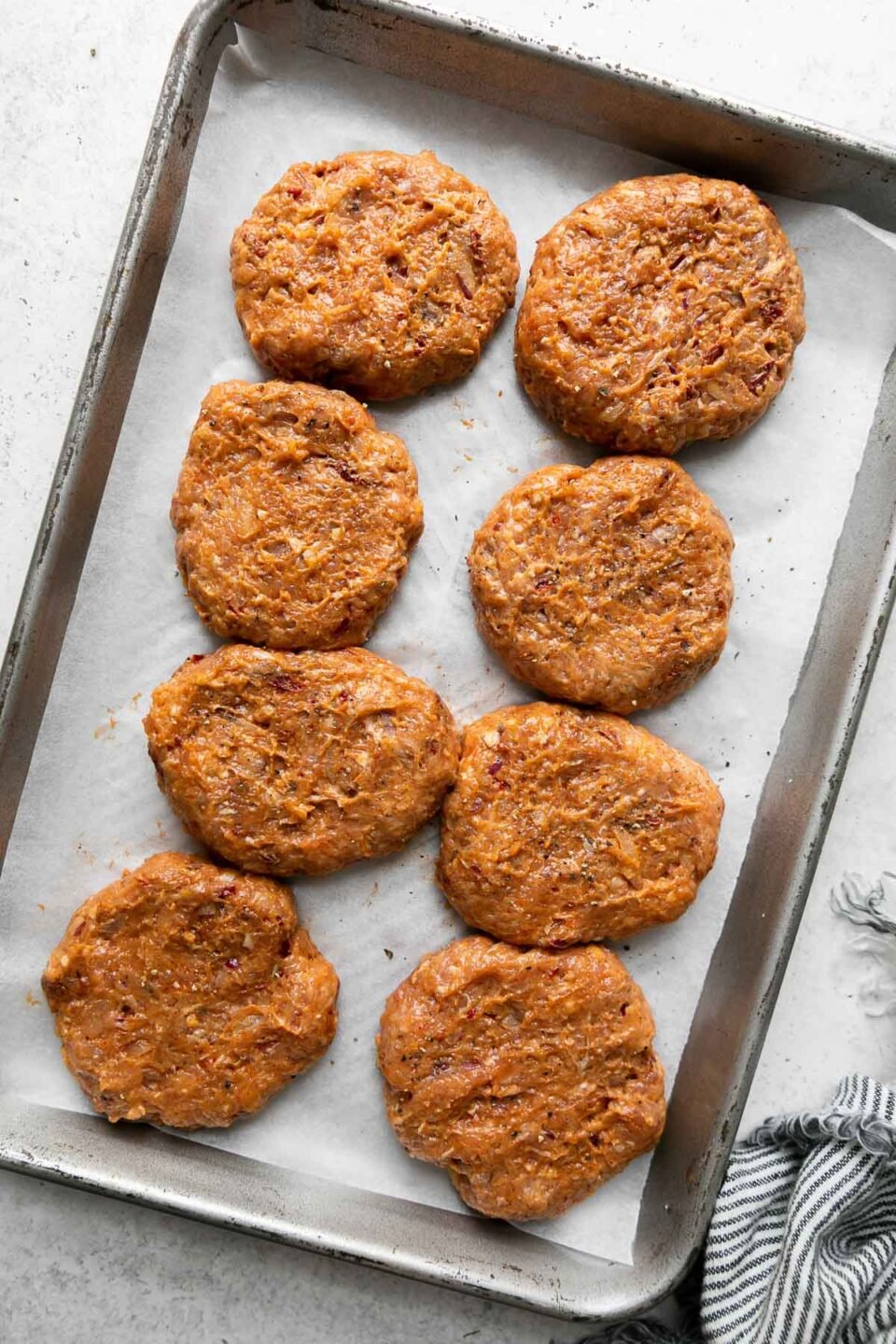 Six formed chipotle turkey burger patties arranged on a parchment paper lined baking sheet. The baking sheet sits atop a creamy white textured surface with a blue and white striped linen napkin resting alongside.