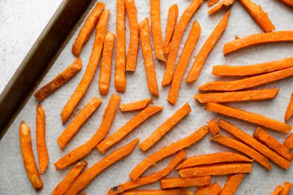 Alexia Sweet Potato Fries with Sea Salt arranged in a single layer on a parchment paper lined baking sheet. The baking sheet sits atop a creamy white textured surface.