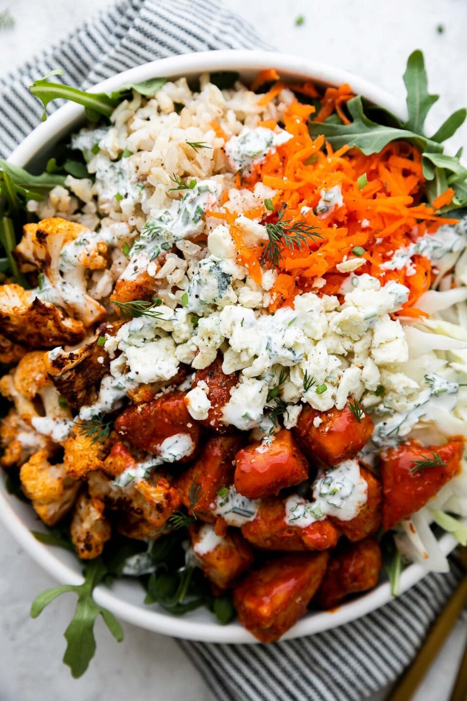 An overhead shot of an assembled & finished Hearty Buffalo Chicken bowl. The bowl is made with cooked brown rice, a bed of arugula, finely shredded cabbage, easy skillet buffalo chicken, roasted cauliflower, grated carrots, cheese, and herby ranch yogurt drizzle over top. The bowls sits atop a creamy white textured surface with a blue and white striped linen napkin resting underneath. Two gold forks rest alongside the bowl.