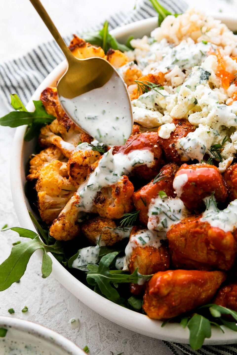 A side angle shot of an assembled & finished Hearty Buffalo Chicken bowl. The bowl is made with cooked brown rice, a bed of arugula, finely shredded cabbage, easy skillet buffalo chicken, roasted cauliflower, grated carrots, cheese, and herby ranch yogurt drizzle over top. The bowls sits atop a creamy white textured surface with a blue and white striped linen napkin resting underneath. A small bowl filled with herby ranch yogurt drizzle sits alongside the buffalo chicken rice bowl and a gold spoon hovers over the buffalo chicken bowl as it is used to drizzle on the ranch yogurt dressing.