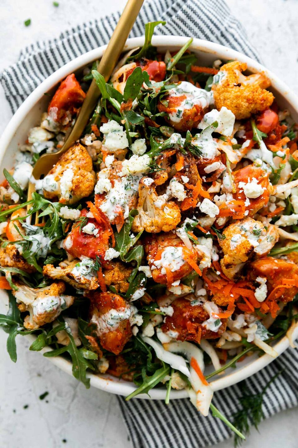 An overhead shot of an assembled & finished Hearty Buffalo Chicken bowl. The bowl is made with cooked brown rice, a bed of arugula, finely shredded cabbage, easy skillet buffalo chicken, roasted cauliflower, grated carrots, cheese, and herby ranch yogurt drizzle over top. The bowls sits atop a creamy white textured surface with a blue and white striped linen napkin resting underneath. A gold fork rests inside of the buffalo chicken rice bowl.