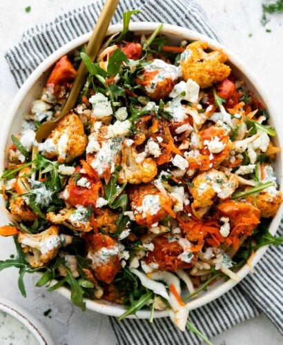 An overhead shot of an assembled & finished Hearty Buffalo Chicken bowl. The bowl is made with cooked brown rice, a bed of arugula, finely shredded cabbage, easy skillet buffalo chicken, roasted cauliflower, grated carrots, cheese, and herby ranch yogurt drizzle over top. The bowls sits atop a creamy white textured surface with a blue and white striped linen napkin resting underneath. A small ceramic bowl filled with herby ranch yogurt drizzle sits alongside the bowl and a gold fork rests inside of the buffalo chicken rice bowl.