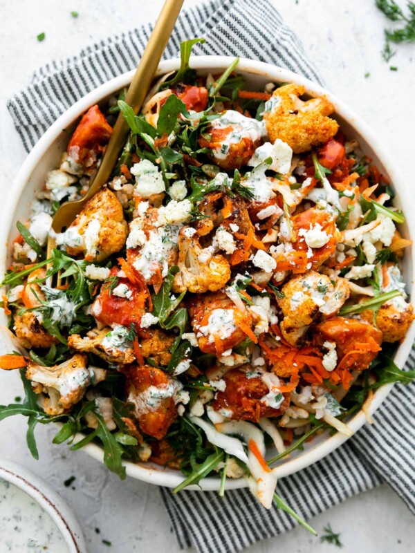 An overhead shot of an assembled & finished Hearty Buffalo Chicken bowl. The bowl is made with cooked brown rice, a bed of arugula, finely shredded cabbage, easy skillet buffalo chicken, roasted cauliflower, grated carrots, cheese, and herby ranch yogurt drizzle over top. The bowls sits atop a creamy white textured surface with a blue and white striped linen napkin resting underneath. A small ceramic bowl filled with herby ranch yogurt drizzle sits alongside the bowl and a gold fork rests inside of the buffalo chicken rice bowl.