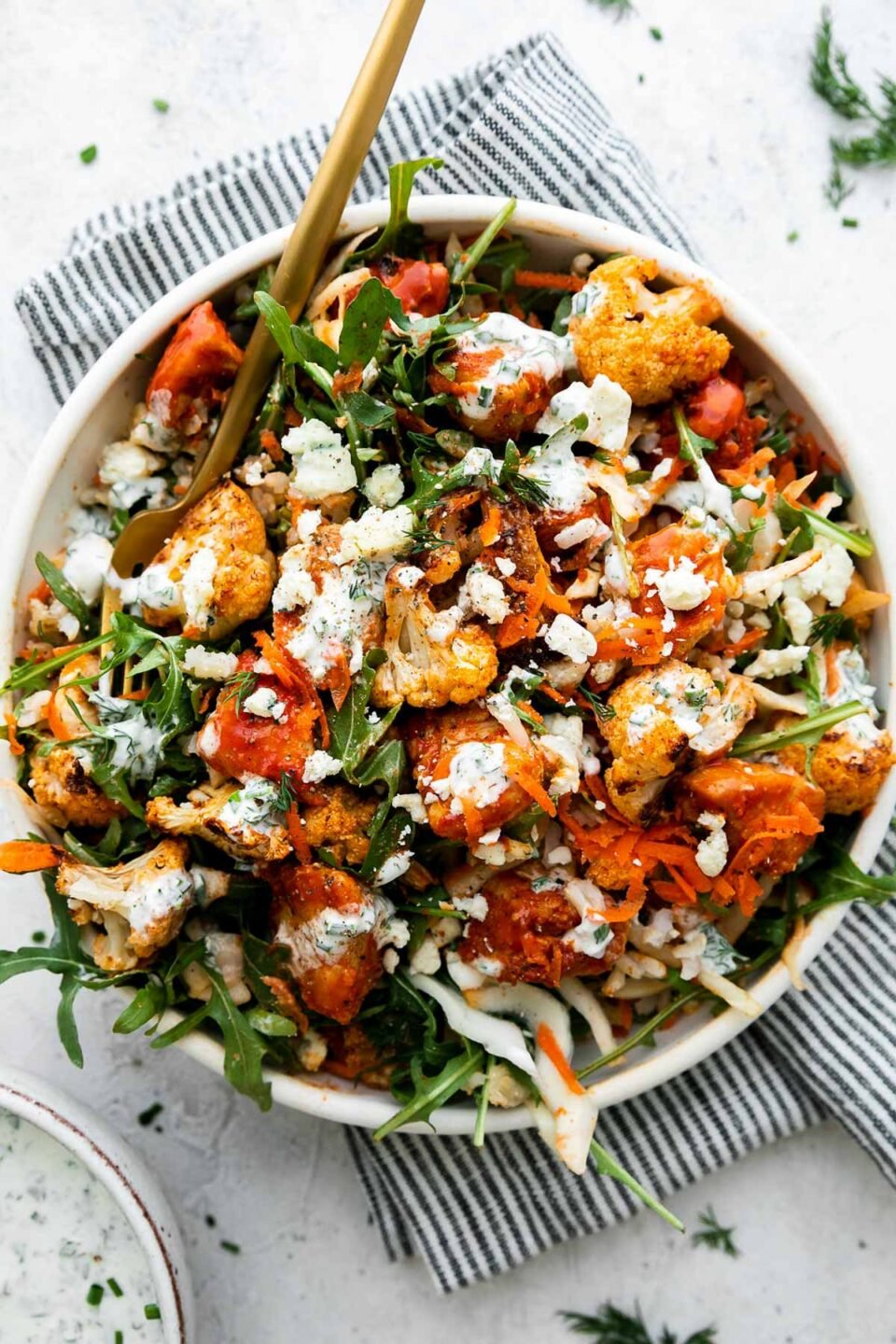 An overhead shot of an assembled & finished Hearty Buffalo Chicken bowl. The bowl is made with cooked brown rice, a bed of arugula, finely shredded cabbage, easy skillet buffalo chicken, roasted cauliflower, grated carrots, cheese, and herby ranch yogurt drizzle over top. The bowls sits atop a creamy white textured surface with a blue and white striped linen napkin resting underneath. A small ceramic bowl filled with herby ranch yogurt drizzle sits alongside the bowl and a gold fork rests inside of the buffalo chicken rice bowl.