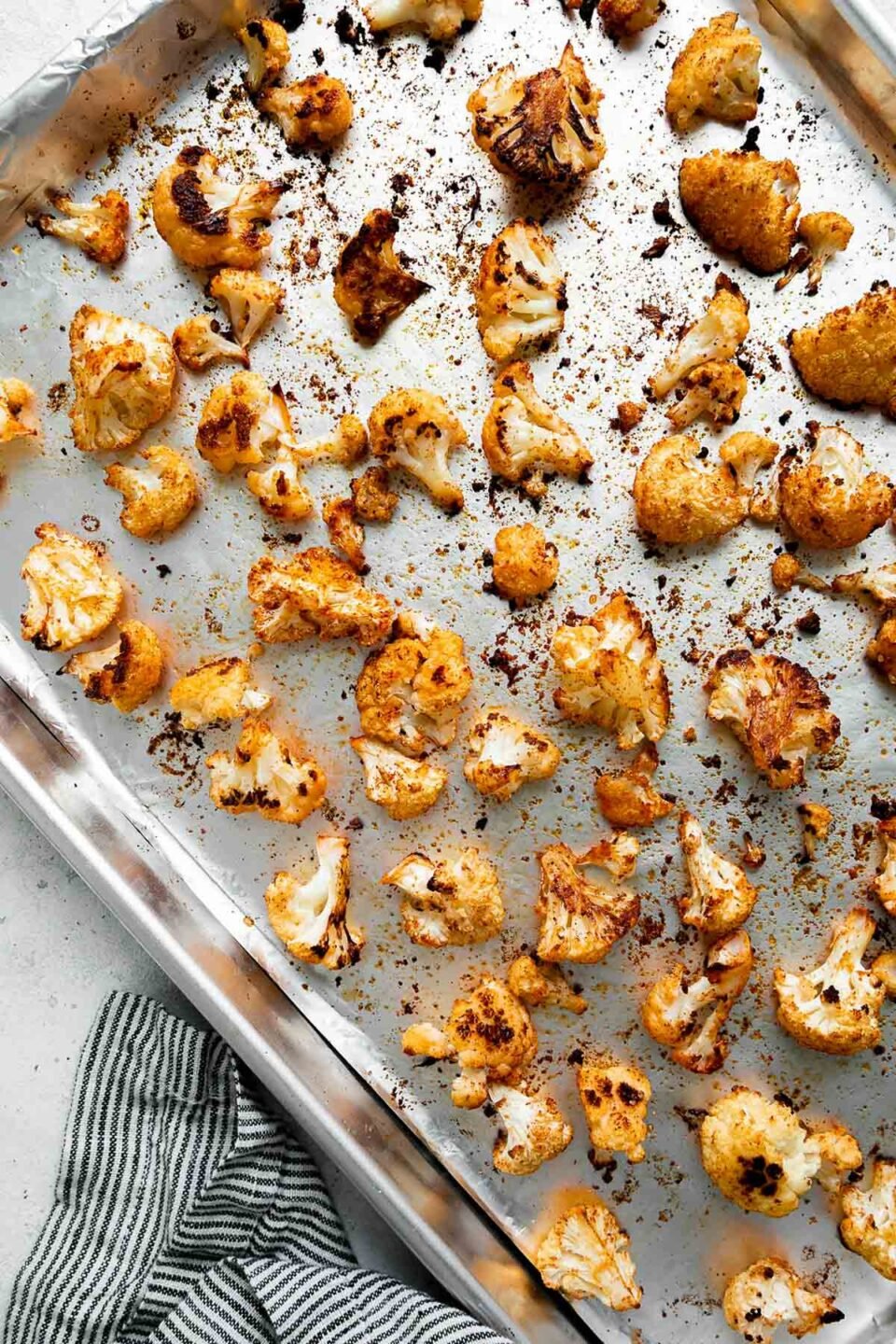 An overhead shot of how to make Hearty Buffalo Chicken Bowls, step 2: Roast the cauliflower. Roasted cauliflower arranged atop an aluminum foil lined baking sheet in a single layer. The cauliflower is roasted until golden brown. The baking sheet sits atop a creamy white textured surface with a blue and white striped linen napkin resting alongside.