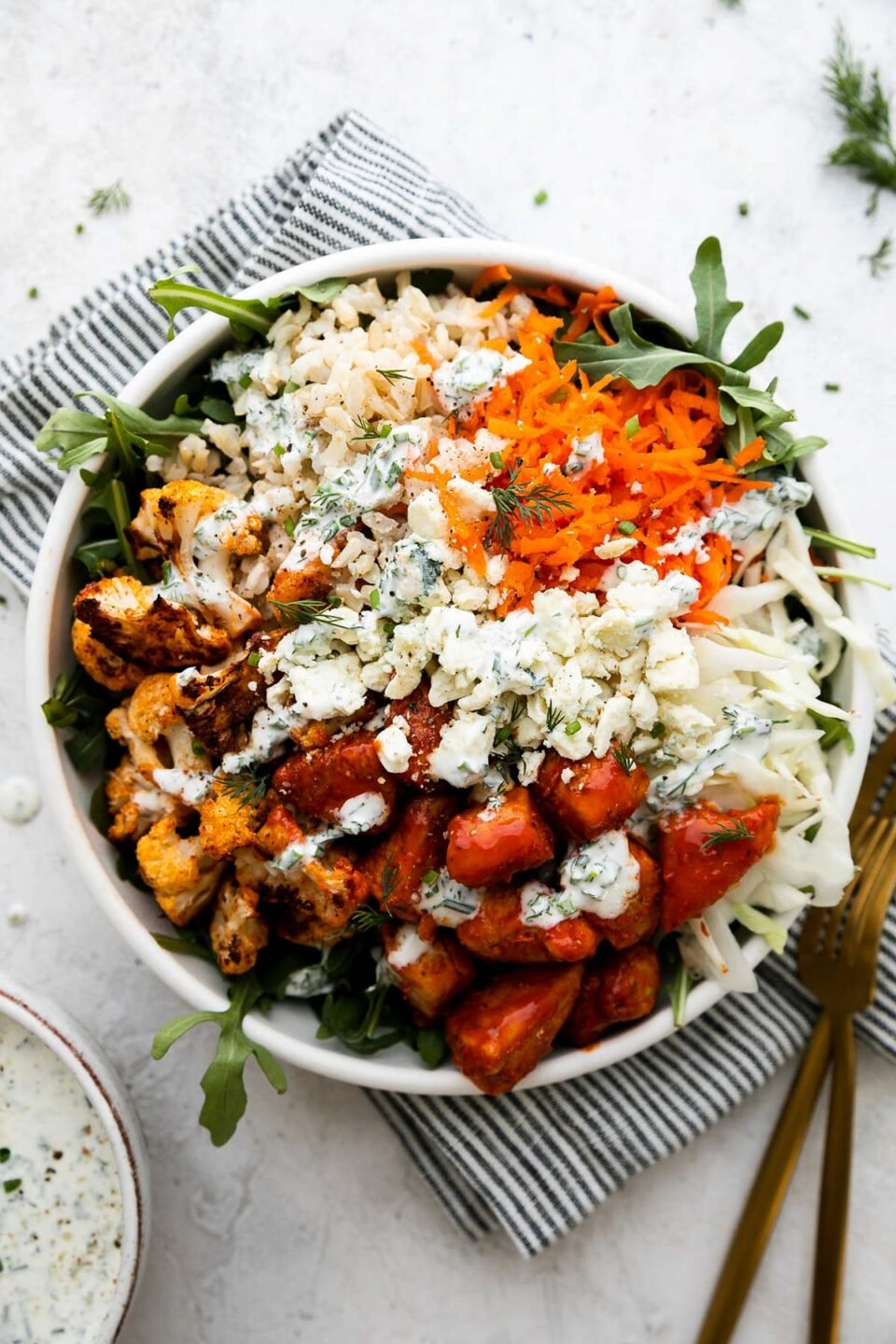 An overhead shot of an assembled & finished Hearty Buffalo Chicken bowl. The bowl is made with cooked brown rice, a bed of arugula, finely shredded cabbage, easy skillet buffalo chicken, roasted cauliflower, grated carrots, cheese, and herby ranch yogurt drizzle over top. The bowls sits atop a creamy white textured surface with a blue and white striped linen napkin resting underneath. Two gold forks, a small ceramic bowl filled with herby ranch yogurt drizzle with a gold spoon resting inside the bowl, freshly snipped chives, and loose fresh dill surround the bowl.