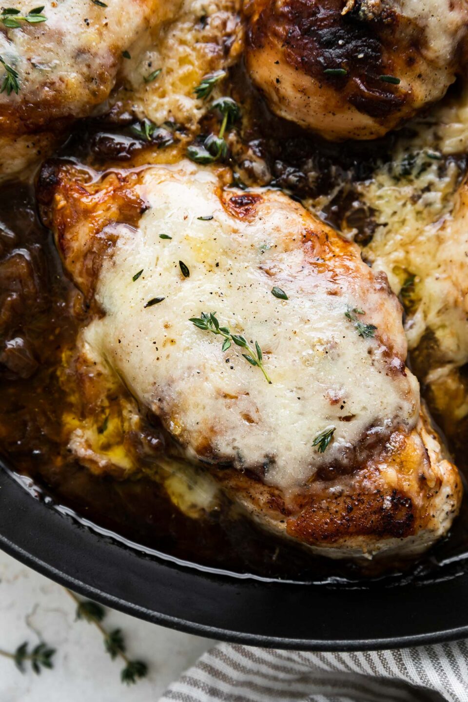 A close up of finished French onion chicken inside of a grey Staub cast iron skillet. The skillet sits atop a creamy white textured surface and has a grey and white striped linen napkin wrapped around the skillet handle. Fresh thyme leaves surround the skillet and the French onion chicken skillet has been garnished with fresh thyme.