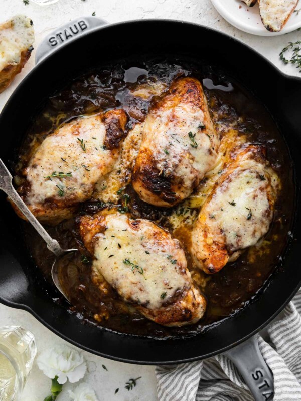Finished French onion chicken inside of a grey Staub cast iron skillet. The skillet ​sits atop a creamy white textured surface and is surrounded by a white and grey striped linen napkin wrapped around the skillet handle, a small white plate of cheesy toasts, a single cheesy toast, white carnation flowers, and two clear glasses filled with white wine. A silver spoon rests inside of the skillet for serving.