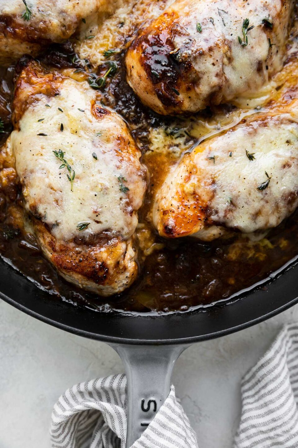 A close up of finished French onion chicken inside of a grey Staub cast iron skillet. The skillet sits atop a creamy white textured surface and has a grey and white striped linen napkin wrapped around the skillet handle.