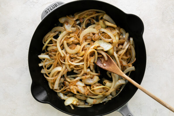 How to make French onion chicken, step 3: Soften the onions. Sliced yellow onion softens inside of a cast iron skillet in melted better. A wooden spoon rests inside of the skillet and the skillet ​sits atop a creamy white textured surface