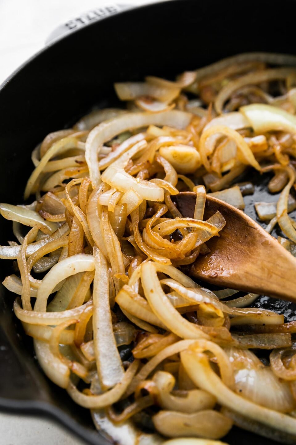 A close up and side angle shot of sliced yellow onion softening inside of a cast iron skillet in melted better. A wooden spoon rests inside of the skillet and the skillet ​sits atop a creamy white textured surface