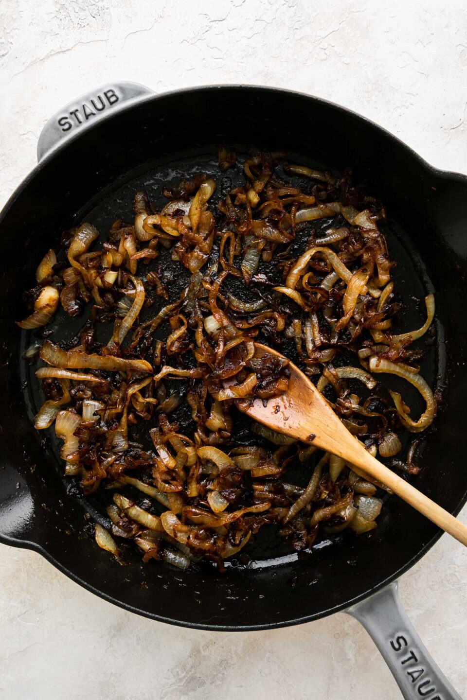 How to make French onion chicken, step 4: Caramelize the onions. Caramelized yellow onions sits inside of a grey Staub cast iron skillet. ​A wooden spoon rests inside of the skillet and the skillet ​sits atop a creamy white textured surface