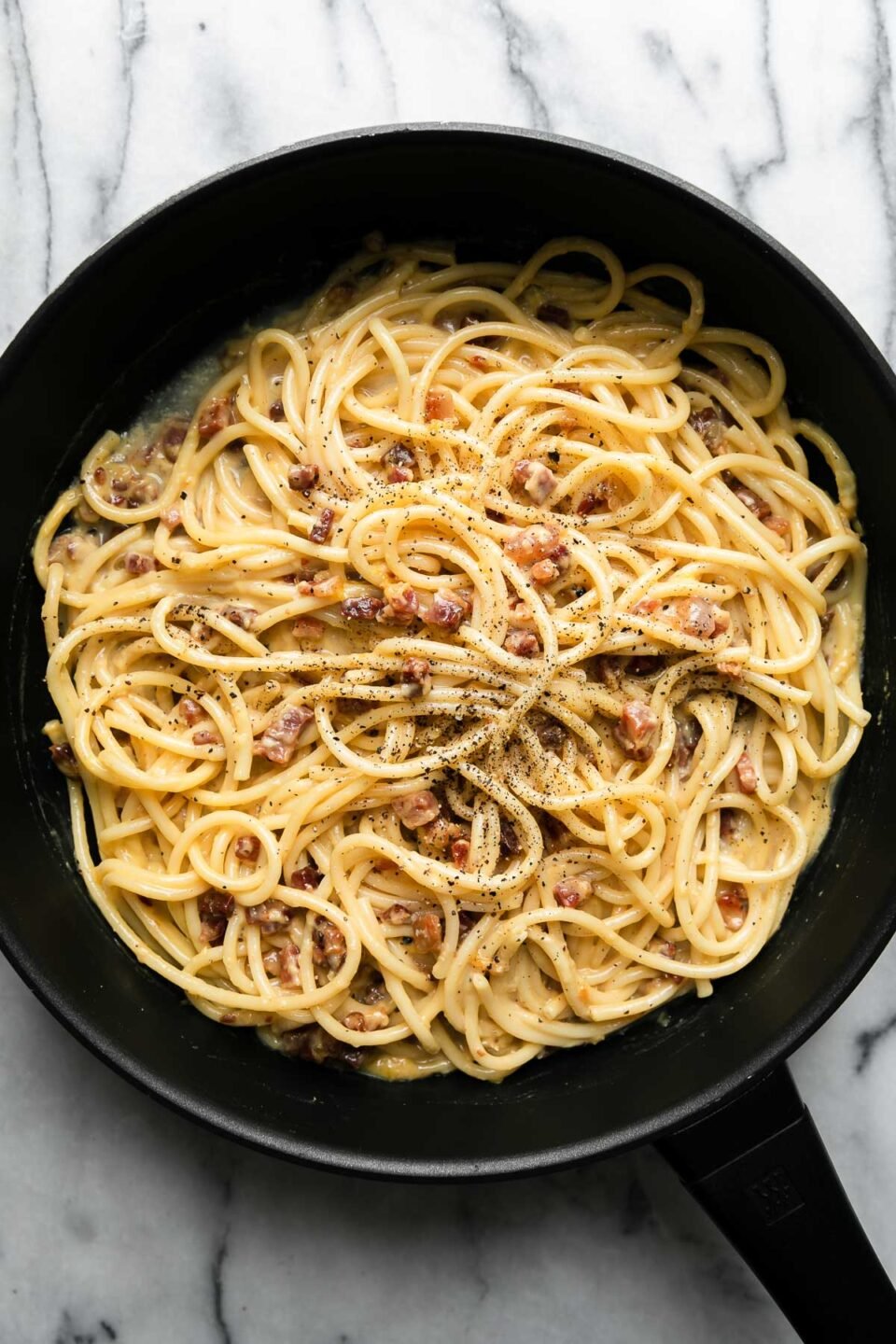 Finished pasta carbonara in a black non-stick skillet garnished with ground black pepper. The skillet sits atop a gray and white marble surface.