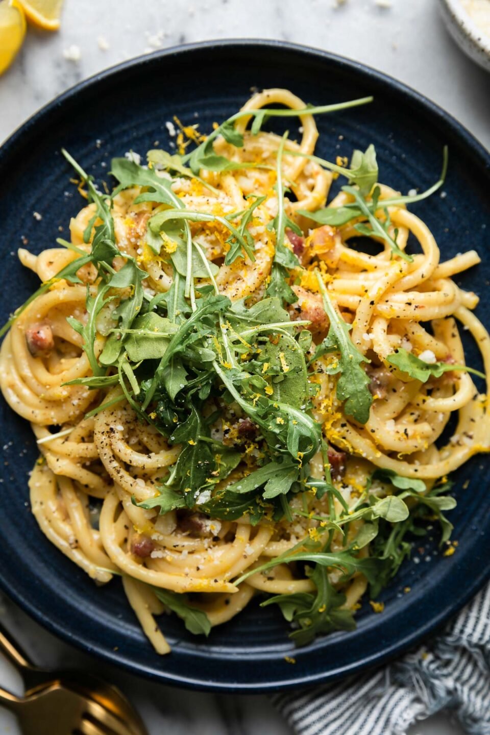 A dark blue ceramic plate topped with bucatini carbonara garnished with ground black pepper, fresh lemon zest, arugula, and freshly grated parmesan cheese sits atop a gray and white marble surface. The plate rests atop a blue and white striped linen napkin with a gold spoon and fork resting alongside. Fresh lemon wedges as well as loose grated parmesan and a small speckled ceramic bowl filled with fresh grated parmesan rest on the surface near the plate.