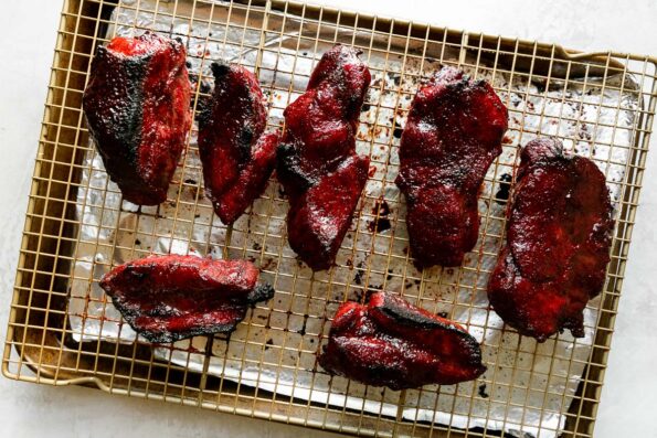 How to make Chinese BBQ pork, step 5: Finish the char siu pork under the broiler. An aluminum foil lined baking sheet sits atop a creamy white textured surface. A wire rack sits atop the baking sheet with pieces of roasted then broiled caramelized char siu pork atop the rack.