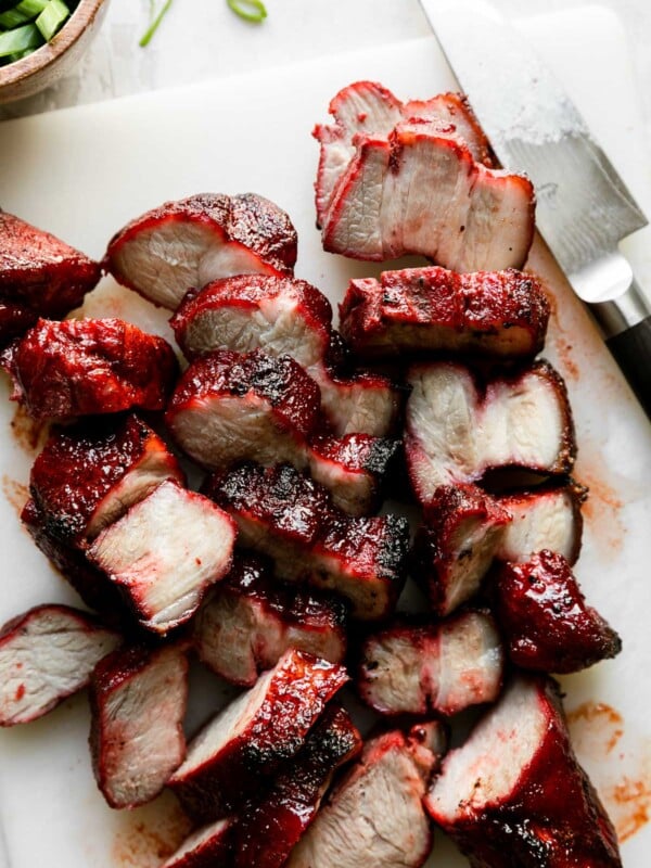 Finished Char Siu pork rests atop a white plastic cutting board atop a creamy white textured surface. The Chinese BBQ pork has been cut into 1/2-inch thick pieces with a chef's knife that sits alongside on the cutting board. A small wooden pinch bowl filled with green onions rests alongside the cutting board.