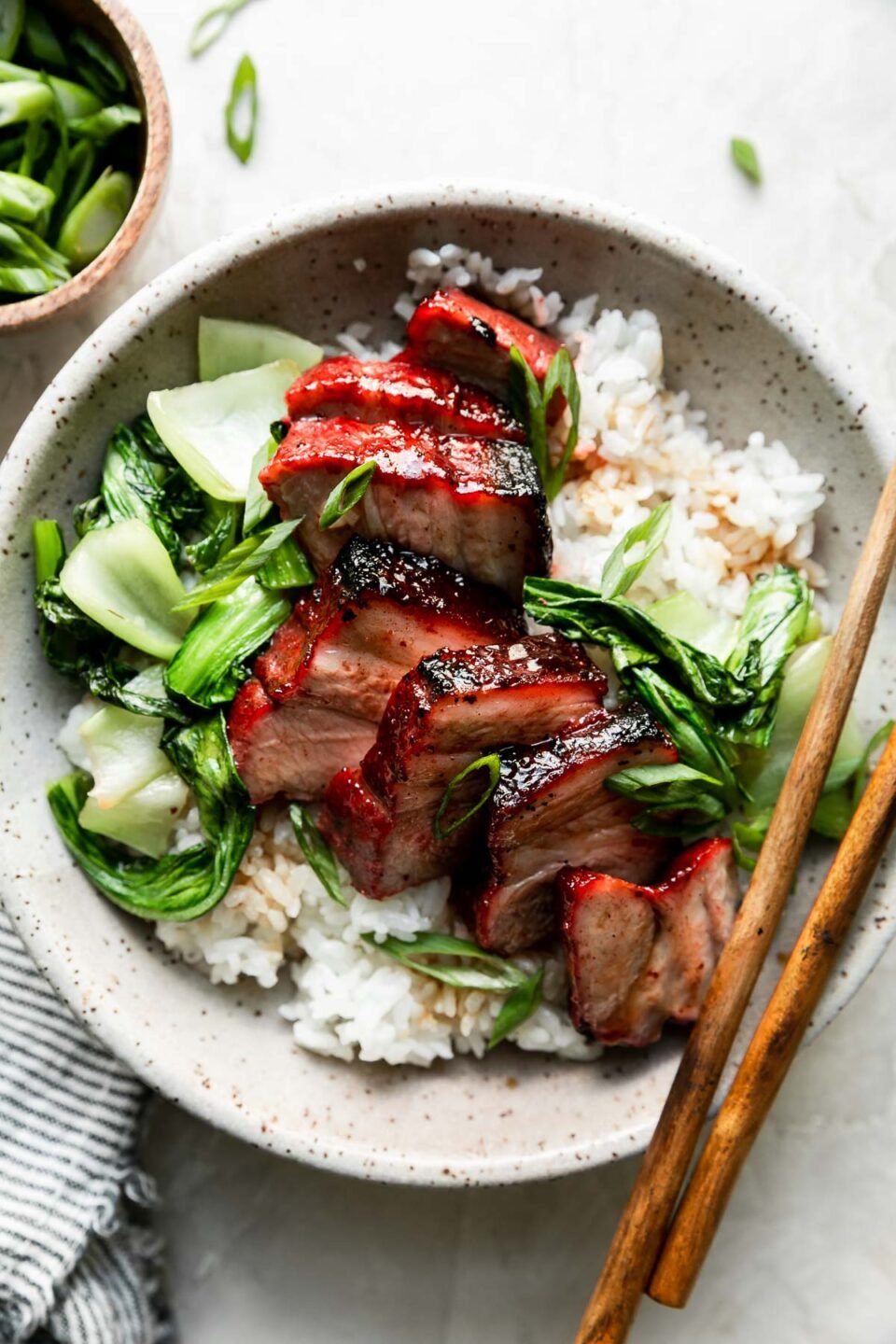 A bowl of char siu pork served over white rice with bok choy & green onions sits atop a creamy white textured surface. Two wooden chop sticks rest atop the bowl. A small wooden pinch bowl filled with sliced green onion and a white and blue striped linen napkin surround the bowl at center.