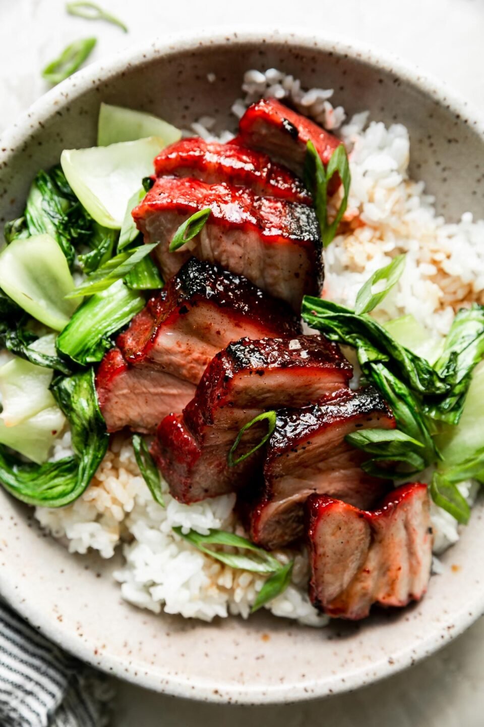 A bowl of char siu pork served over white rice with bok choy & green onions sits atop a creamy white textured surface. Loose pieces of sliced green onion and a white and blue striped linen napkin surround the bowl at center.