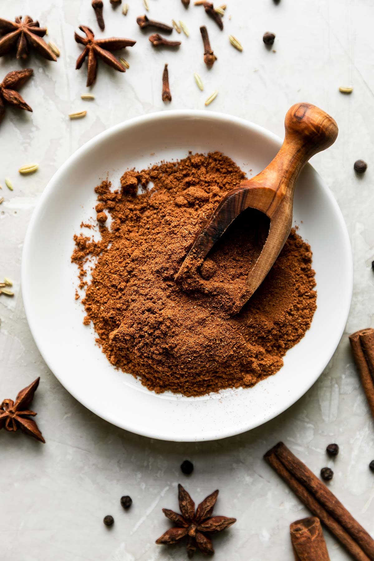 Ground Chinese five spice in a white shallow bowl atop a creamy white textured surface. A wooden scoop rests inside of the ground Chinese five spice with whole spices: cinnamon, cloves, fennel, & star anise surrounding.