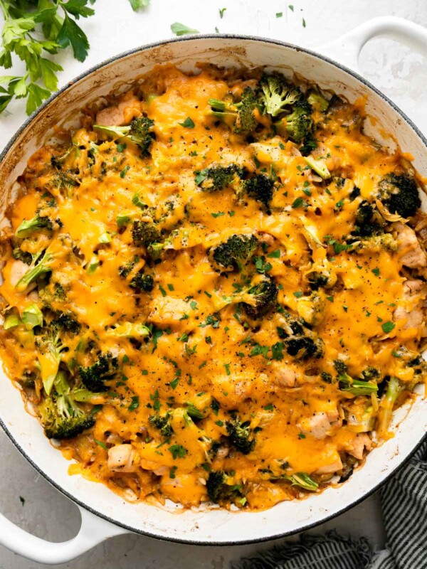 Finished cheesy chicken and broccoli orzo skillet fills a white double-handle skillet. The skillet sits atop a creamy white textured surface and is surrounded by fresh parsley and a blue and white striped linen napkin. The skillet dinner has been garnished with chopped fresh parsley and ground black pepper.
