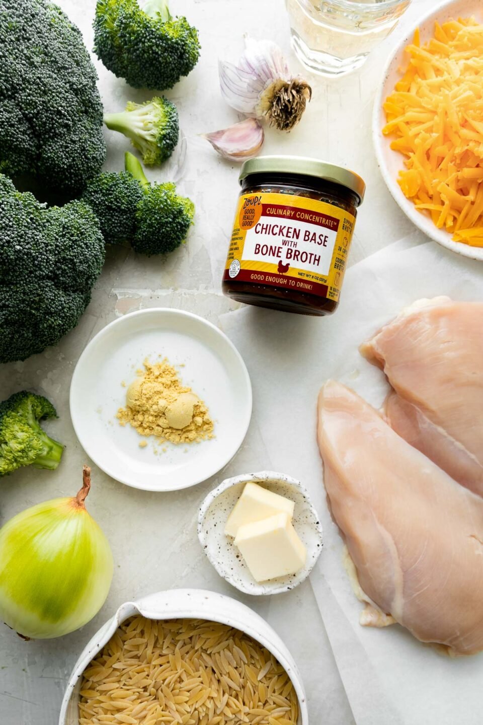 Cheesy Chicken and Broccoli Orzo Skillet ingredients arranged on a creamy white textured surface: unsalted butter, boneless, skinless chicken breasts, yellow onion, garlic, mustard powder, orzo, dry white wine, Zoup! Chicken Bone Broth Concentrate, broccoli, and sharp cheddar cheese.
