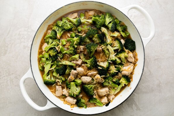 How to make chicken and broccoli orzo, step 5: Build the chicken and broccoli orzo skillet. Broccoli florets and browned chicken are added to toasted orzo pasta and cooked aromatics in a white double-handle skillet. The skillet sits atop a creamy white textured surface.
