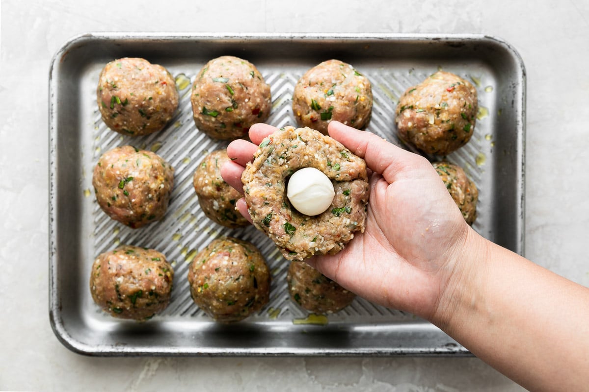 How to make Mozzarella Stuffed Chicken Parmesan Meatballs, step 3: Form & stuff the chicken parmesan meatballs. 11 formed Chicken parmesan meatballs are arranged in rows of three on a small aluminum baking sheet. A woman's hand holds a single flat meatball patty with a fresh mozzarella ball placed at center above the baking sheet. The baking sheet sits atop a creamy white textured surface.