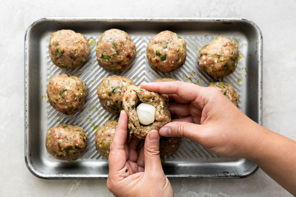 How to make Mozzarella Stuffed Chicken Parmesan Meatballs, step 3: Form & stuff the chicken parmesan meatballs. 11 formed Chicken parmesan meatballs are arranged in rows of three on a small aluminum baking sheet. A woman's hands work to wrap the edges of a flat meatball patty around a ball of fresh mozzarella to create a mozzarella stuffed chicken meatball. The baking sheet sits atop a creamy white textured surface.
