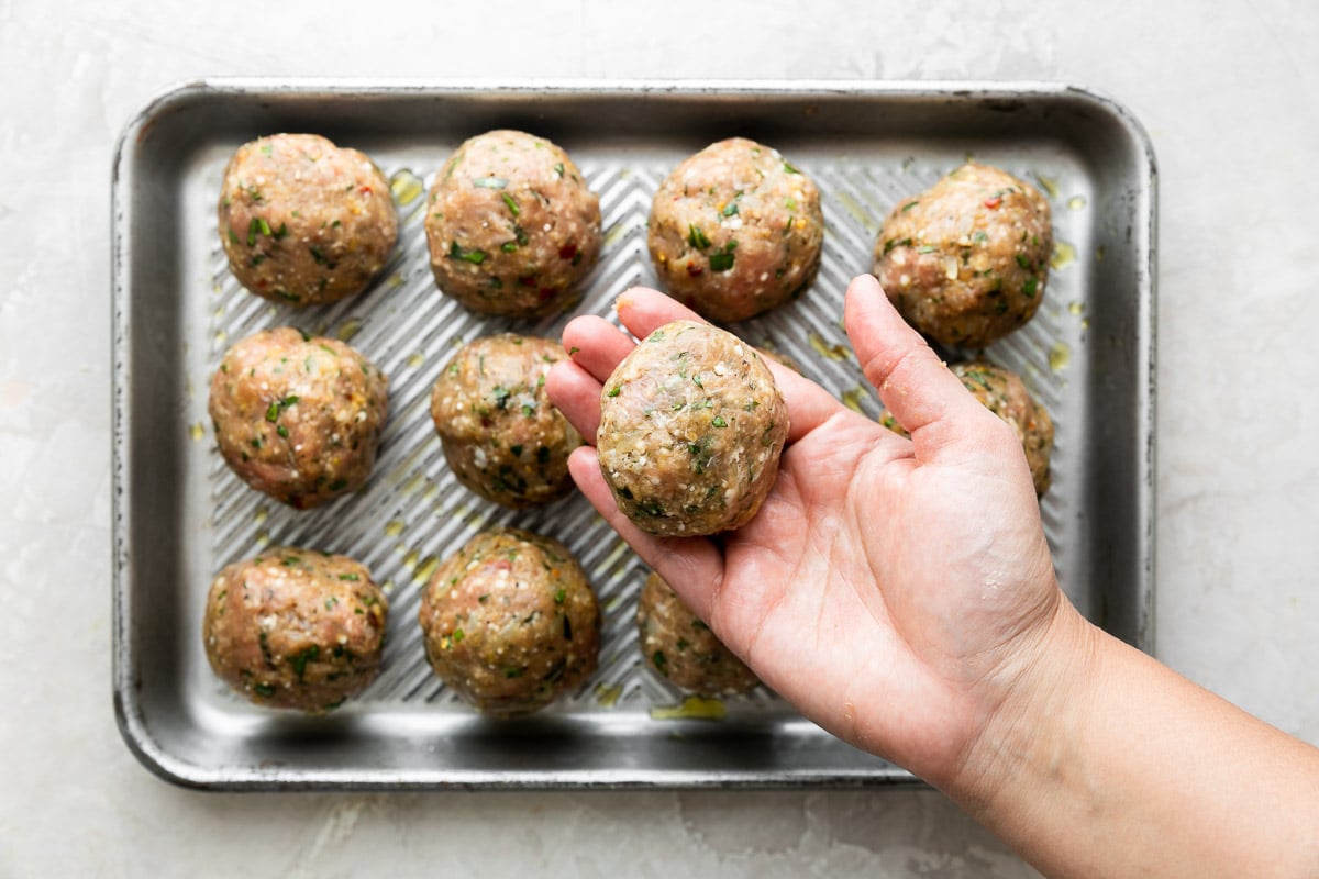 How to make Mozzarella Stuffed Chicken Parmesan Meatballs, step 3: Form & stuff the chicken parmesan meatballs. 11 formed Chicken parmesan meatballs are arranged in rows of three on a small aluminum baking sheet. A woman's hands finish closing up the the edges of meatballs mixture that engulfs a ball of fresh mozzarella to create a mozzarella stuffed chicken meatball. The baking sheet sits atop a creamy white textured surface.