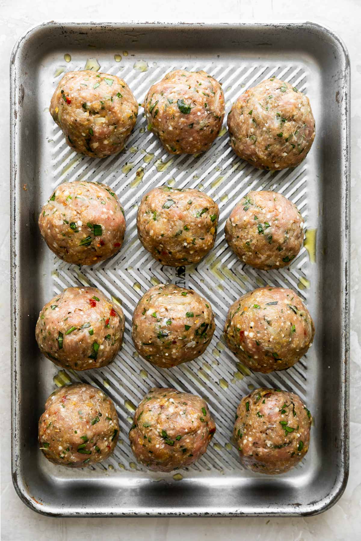 Twelve formed but uncooked Chicken parmesan meatballs are arranged in rows of three on a small aluminum baking sheet. The baking sheet sits atop a creamy white textured surface.