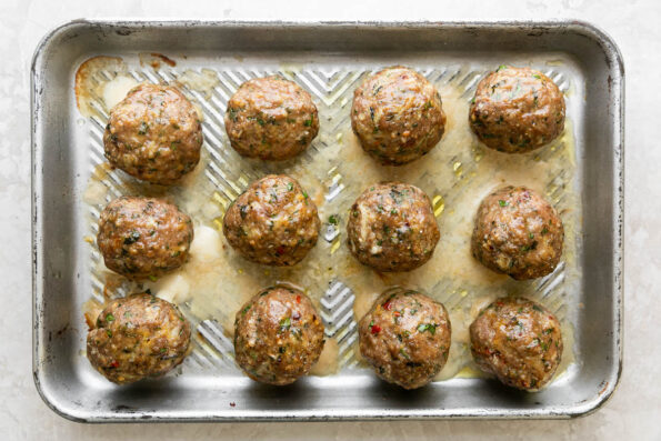 Twelve cooked Chicken Parmesan Meatballs are arranged in rows of three on a small aluminum baking sheet. The baking sheet sits atop a creamy white textured surface.