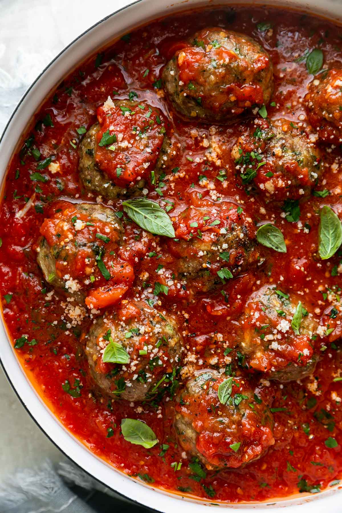 Mozzarella Stuffed Chicken Parmesan Meatballs simmer in a red tomato sauce in a large white skillet. The meatballs have been garnished with chopped basil and grated parmesan and the skillet sits atop a creamy white textured surface with a light blue linen napkin tucked underneath the skillet.