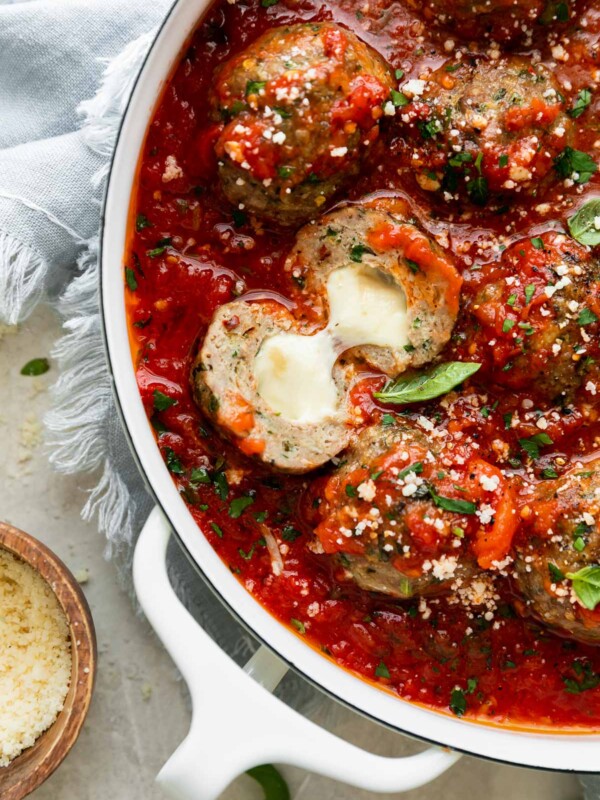 Mozzarella Stuffed Chicken Parmesan Meatballs simmer in a red tomato sauce in a large white double-handled skillet. The meatballs have been garnished with chopped basil and grated parmesan and the skillet sits atop a creamy white textured surface with a light blue linen napkin tucked underneath the skillet. A small wooden pinch bowl filled with grated parmesan and a few loose fresh basil leaves surround the skillet. Inside of the skillet one meatballs has been cut in half and folded open to reveal the mozzarella stuffed interior.