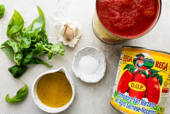 Simple San Marzano Tomato Sauce ingredients arranged on a creamy white textured surface: whole peeled San Marzano tomatoes, extra virgin olive oil, garlic, fresh basil, granulated sugar, kosher salt and ground black pepper, and optional crushed red pepper.