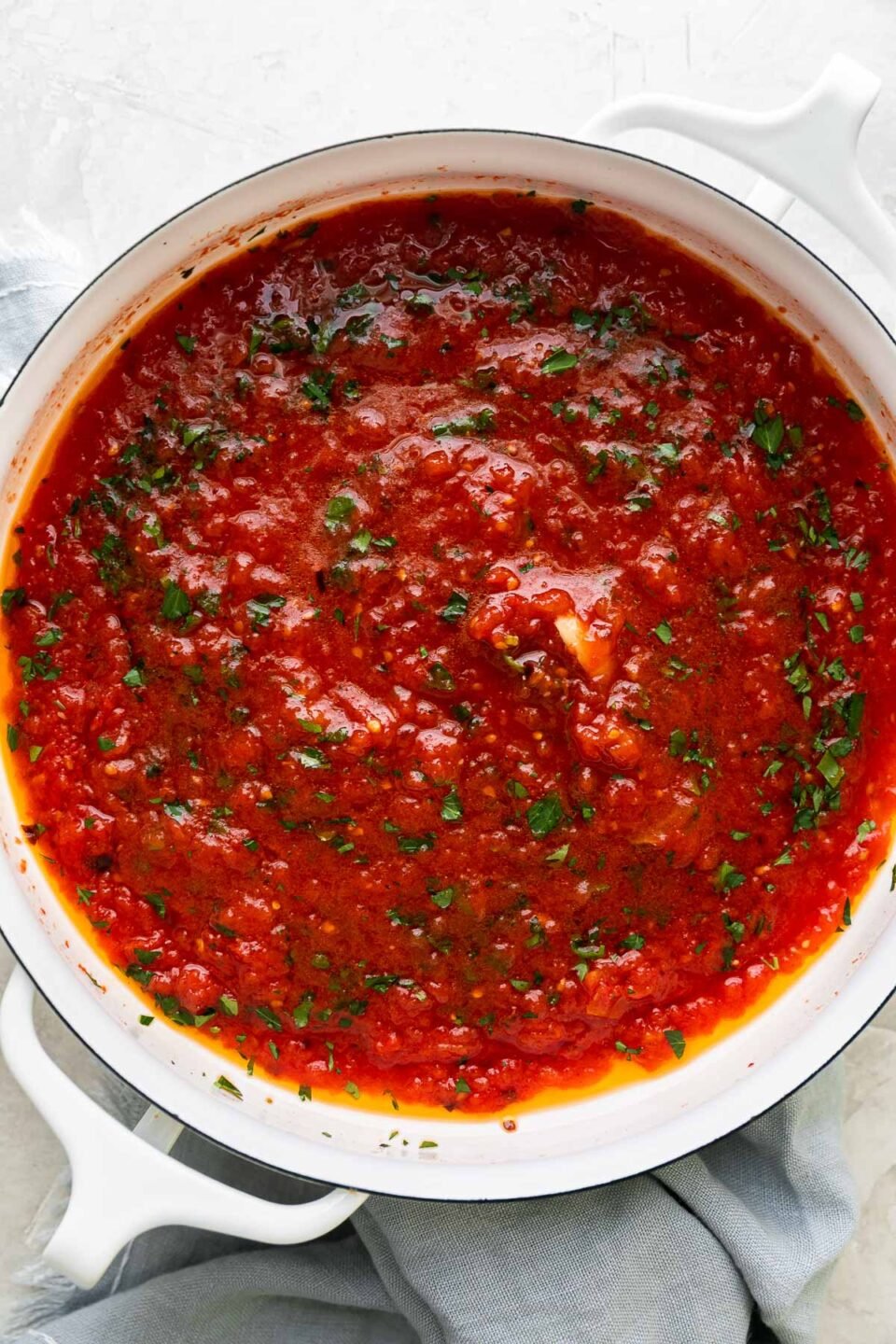 Finished San Marzano tomato sauce fills a large white double-handled skillet. The quick tomato sauce is garnished with chopped basil and the skillet sits atop a creamy white textured surface. A light blue linen napkin rests alongside the skillet.