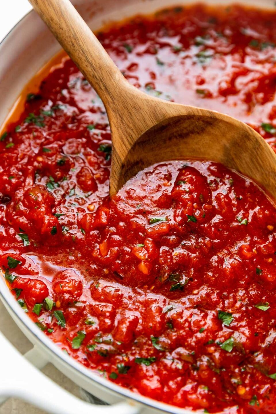 A side angle shot of finished San Marzano tomato sauce fills a large white double-handled skillet. The quick tomato sauce is garnished with chopped basil and the skillet sits atop a creamy white textured surface. A wooden spoon rests inside of the skillet for serving.