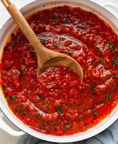 An overhead shot of finished San Marzano tomato sauce fills a large white double-handled skillet. The quick tomato sauce is garnished with chopped basil and the skillet sits atop a creamy white textured surface. A wooden spoon rests inside of the skillet for serving and a light blue linen napkin is tucked underneath the skillet.