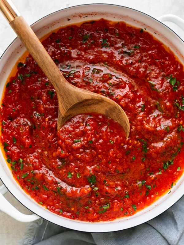 An overhead shot of tomato sauce in a large white double-handled skillet. The sauce is garnished with chopped basil and the skillet sits atop a creamy white textured surface. A wooden spoon rests inside of the skillet for serving and a light blue linen napkin is tucked underneath the skillet.