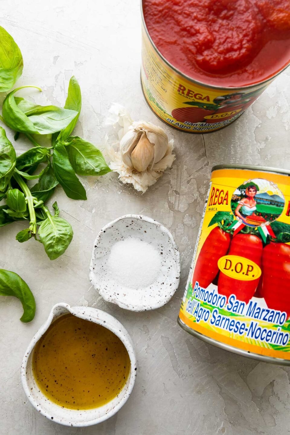 Simple San Marzano Tomato Sauce ingredients arranged on a creamy white textured surface: whole peeled San Marzano tomatoes, extra virgin olive oil, garlic, fresh basil, granulated sugar, kosher salt and ground black pepper, and optional crushed red pepper.
