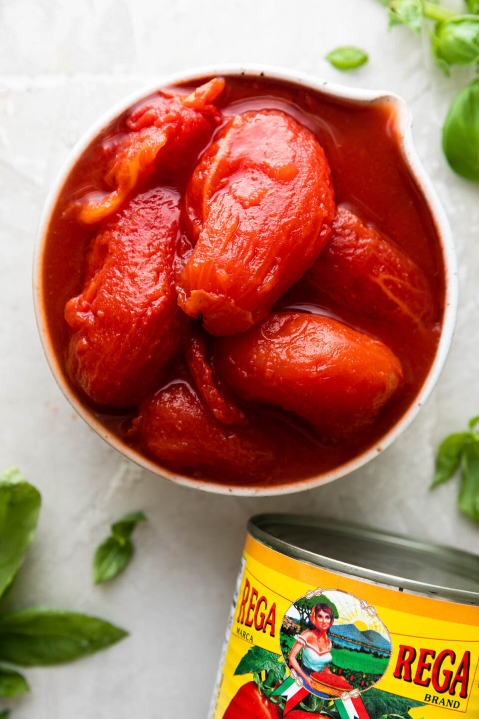 Whole peeled San Marzano tomatoes for a simple tomato sauce fill a small white ceramic bowl. The bowl sits atop a creamy white textured surface. A can of unopened San Marzano tomatoes rests alongside the bowl with the label facing upwards and loose fresh basil leaves surround the bowl.