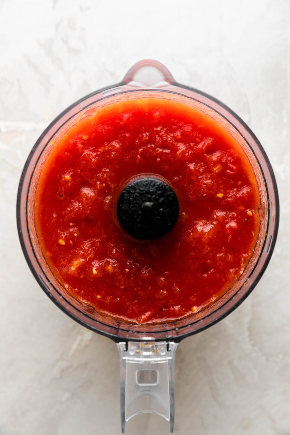 An overhead shot of a food processor bowl is filled with tomatoes that have been pulsed and chopped leaving some textured to use in a quick tomato sauce. The food processor bowl sits atop a creamy white surface.