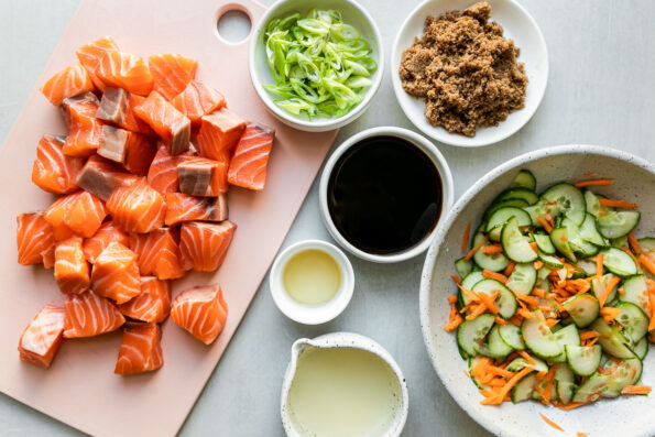 Soy-glazed Seared Salmon rice bowl ingredients arranged atop a gray surface: prepared cucumber & carrot namasu, fresh wild-caught salmon, green onions, brown sugar, soy sauce, mirin, grapeseed oil.