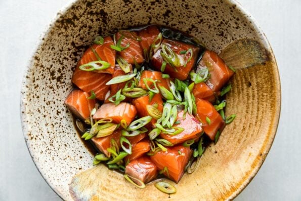 How to make a rice bowl with salmon, step 2: Marinate the salmon. Cubed fresh wild-caught salmon marinates in brown sugar, soy sauce, and mirin within a small speckled ceramic bowl with slices of green onion. The bowl sits atop a light gray surface.