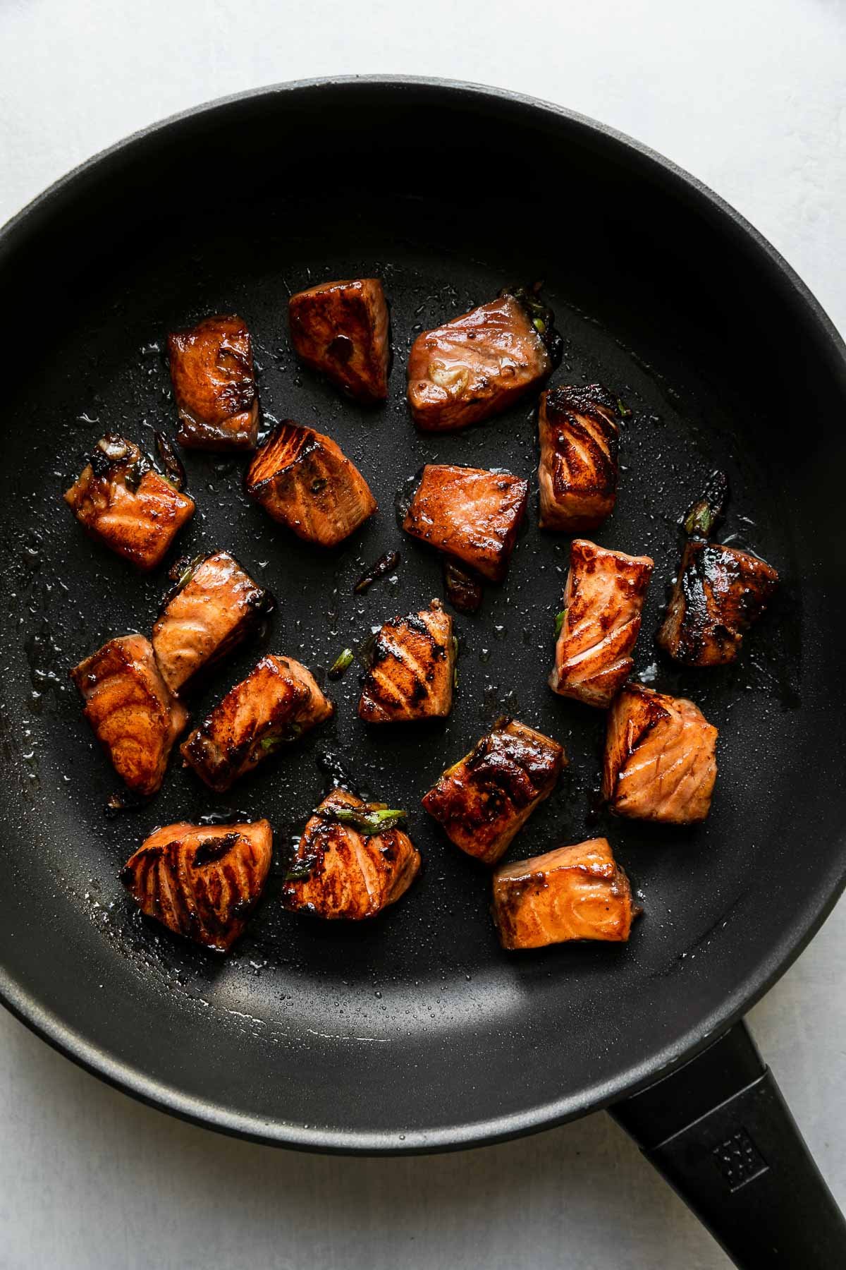 How to make a salmon and rice bowl, step 3: Sear the salmon. Cubed marinated salmon sears inside of a black non-stick frying pan. The frying pan sits atop a light gray surface.