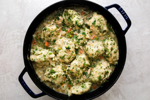 Finished homemade chicken and dumplings fill a dark blue Staub cocotte. The cocotte sits atop a creamy white textured surface.